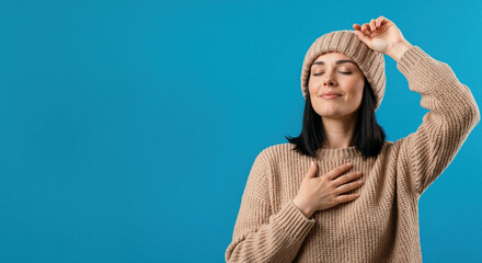 A woman in a cozy winter sweater and beanie feeling grateful. Self-care and comfort concept. Studio portrait isolated on a blue background with copy space
