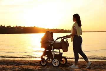 Happy mother with baby in stroller walking near river at sunset