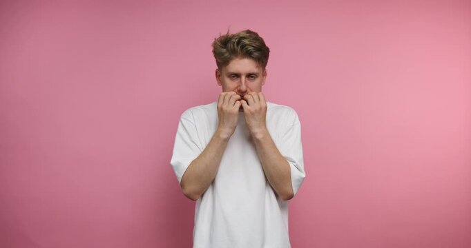 Young man shows different emotions in front of a pink wall while covering his mouth and expressing surprise or fear