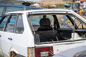 Abandoned white car with shattered windows and rusted body sits in a junkyard among other wrecked vehicles