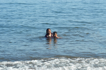 Fototapeta premium A cheerful mother walks toward her young son in the shallow surf, smiling warmly as they enjoy playful water activities together in the gentle morning tide at the beach.