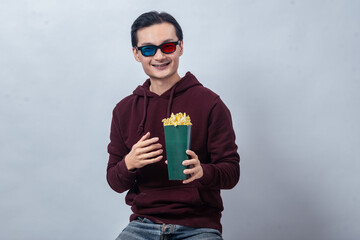 Asian man in maroon hoodie wearing 3D glasses, holding a green container of popcorn and smiling...