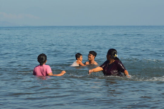 The mother splashes water joyfully to motivate her son swimming toward his father with a ball, as the daughter also enjoys the seaside play. - Powered by Adobe