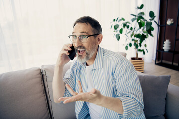 Mature bearded man happily talking on a smartphone while seated indoors, surrounded by a stylish and cozy environment