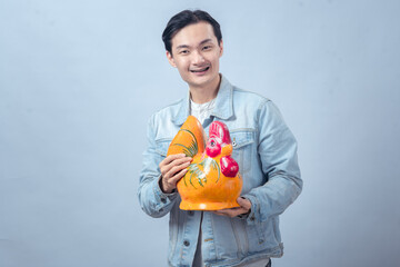 Asian man in a denim jacket smiling while holding a large colorful chicken-shaped ceramic decoration against a plain background