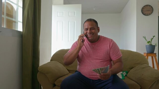 A man sitting on a couch with a cell phone and a stack of money in his hand. He is smiling and he is happy