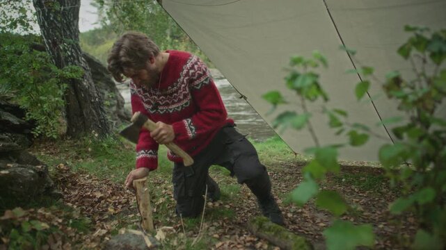 Survivalist chopping wood with hatchet while preparing to make fire near tarp shelter on riverbank in forest