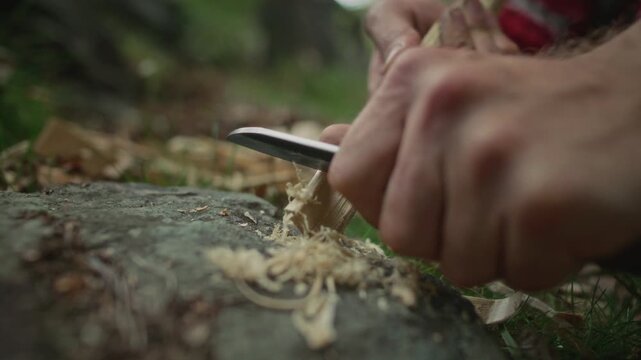 Hands of survivalist making feather sticks with knife on forest floor to start fire in wilderness. Close-up shot