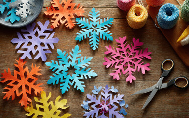Fototapeta premium Colorful paper snowflakes in various shapes and sizes are scattered on a wooden table, surrounded by crafting materials, showcasing a festive and creative atmosphere for winter decorations