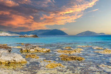 A clear, deep blue sea. In the foreground, rocks and stones are in the water and close to the...