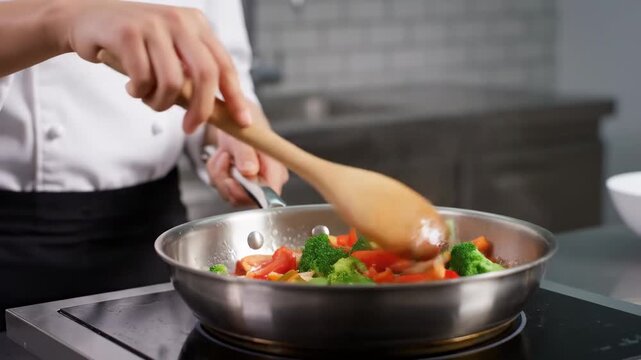 Chef Stirring Colorful Vegetables in Pan - A chef in a white jacket uses a wooden spoon to stir colorful vegetables, including broccoli and carrots, cooking in a stainless steel frying pan on a