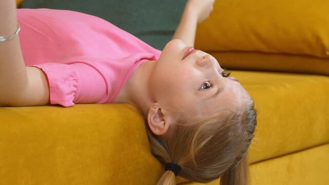 Young teenage girl in pink shirt comfortably seated on mustard-colored sofa, changes posture and looks up with a peaceful, thoughtful expression on her face