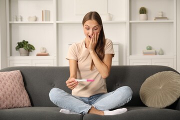 Excited young woman with pregnancy test on sofa at home