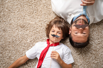 Father and son with paper moustache lying on rug, top view. Space for text