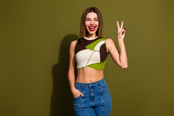Obraz na płótnie Canvas Young fashionable woman making peace sign against olive green background, wearing trendy top and denim jeans, smiling confidently