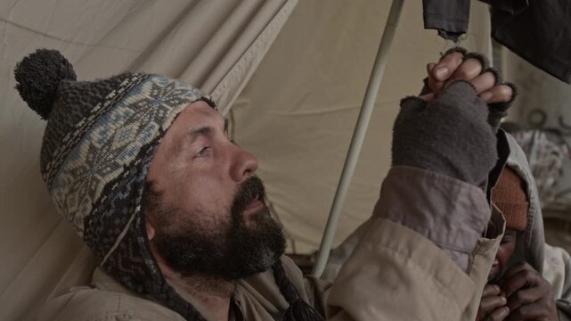 Close-up shot of Caucasian male beggar wearing hat and jacket threading needle while Black vagrant playing harmonica in homeless camp