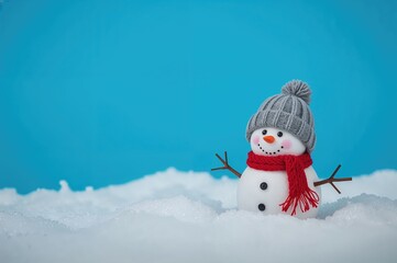Obraz premium How to reproduce a holiday greeting card. A charming little snowman with a knitted hat and scarf stands in the snow against a bright blue backdrop. Amusing snowman with a seasonal setting. Greeting