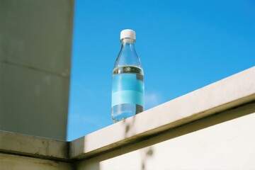 A clear water bottle with a blank blue label rests on a ledge against a bright blue sky, conveying refreshment, simplicity, and everyday hydration.