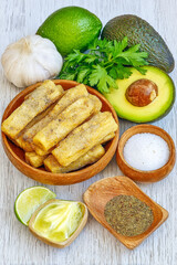 A wooden bowl of seasoned baked yuca sticks served with fresh avocado, lime, garlic, and spices on a white wooden background.