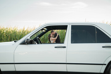 A romantic couple shares a tender moment by a classic white car, holding hands and leaning in for a kiss on a sunny day outdoors.