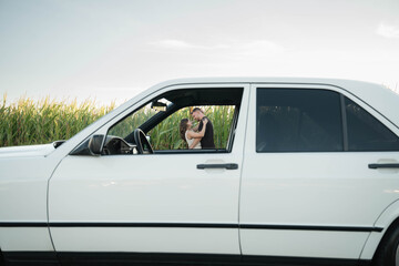 A romantic couple shares a tender moment by a classic white car, holding hands and leaning in for a kiss on a sunny day outdoors.
