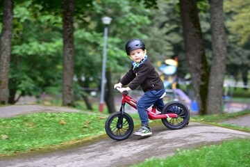 Naklejka premium Young boy riding balance bike with helmet on curved asphalt track. Child enjoys sport activity, balancing carefully while cycling outdoors on pump track in autumn season.