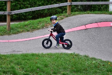 Young boy riding balance bike with helmet on curved asphalt track. Child enjoys sport activity, balancing carefully while cycling outdoors on pump track in autumn season.