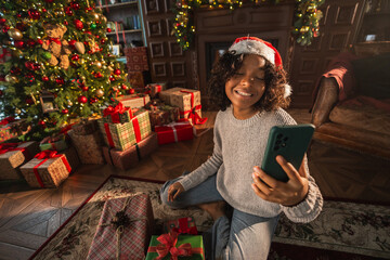 Merry Christmas. African woman in Santa hat having video chat in smartphone. Blogger in festive...
