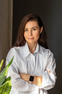 Confident woman with long brown hair stands with crossed arms in room. She wears white shirt and has a friendly expression. Business portrait of designer, businesswoman, successful woman 42 years old