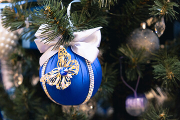 Elegant blue Christmas ball decorated with a shiny butterfly and white satin bow, hanging on a festive tree with sparkling lights and needles