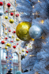 Close-up of white artificial Christmas tree decorated with shiny gold, silver, and red balls on a festive city street with holiday lights and garlands
