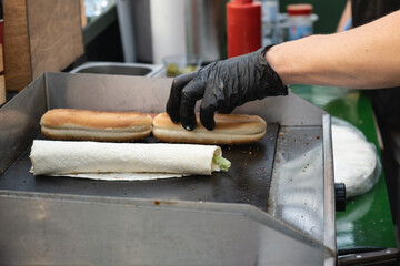 Close-up of a street food vendor in black gloves grilling sandwich buns and a wrap with lettuce on a hot flat-top grill at a local outdoor event