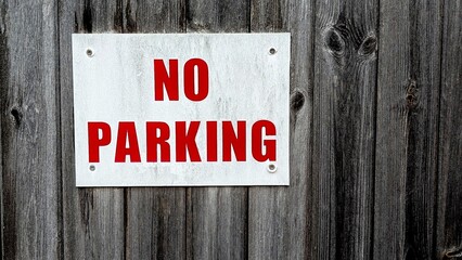 'No Parking' sign with red letters on a white board against a weathered gray wooden fence, ideal...