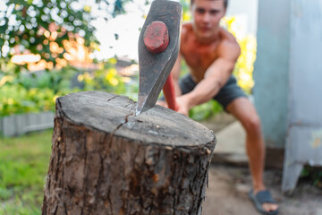 A young tanned muscular man with a bare torso cuts firewood with an axe in his backyard in the summer. Sport and fitness. Muscular body.