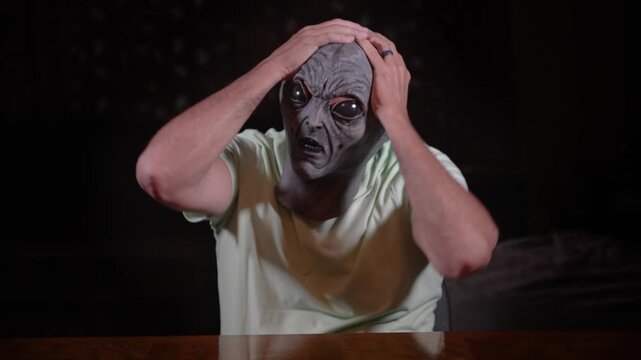A person wearing a detailed alien mask sits at a table, gripping their head with both hands, conveying confusion, frustration, or a sense of giving up. 14826  	