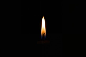 A lone candle shines quietly in the dark, its gentle light spreading warmth around. The flame dances softly, featuring a blue core and a golden peak, creating a subtle glow on the dark backdrop.