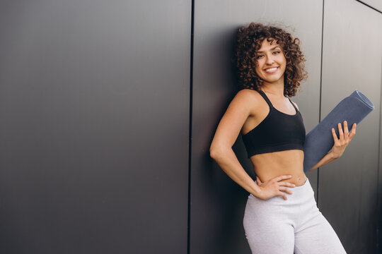 Smiling sportswoman holding yoga mat leaning against gray wall