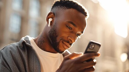 Young handsome stylish man with wireless earbuds using smartphone outdoors in sunlight. Modern lifestyle, mobile technology, digital communication, urban street background, trendy minimal portrait.