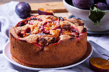 Plum pie with filling and sugar crust, sprinkled with powdered sugar and plums on a wooden table.