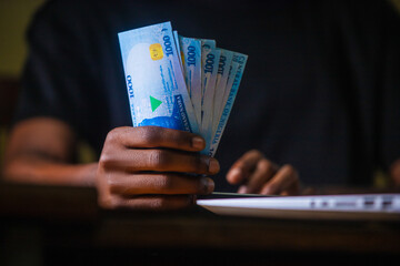 Dark skinned African male wearing black shirt sitting at desk, working on his laptop and holding Nigerian Naira notes