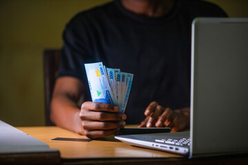 Dark skinned African male wearing black shirt sitting at desk, working on his laptop and holding Nigerian Naira notes