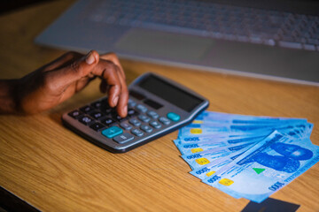 Dark skinned African person sitting punching pressing calculator with Nigerian Naira notes on table desk