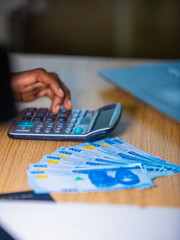 Vertical shot of Dark skinned African person sitting punching pressing calculator with one thousand Nigerian naira notes on table desk