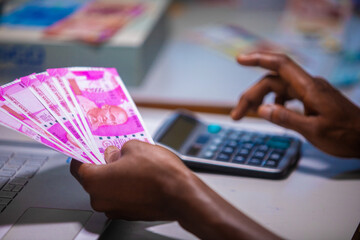 Dark skinned African person sitting punching pressing calculator with Indian Rupee notes on table desk