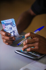 Dark skinned African person working on desk holding Saudi riyal notes in hand and pen pressing punching calculator  on table. accounting concept.
