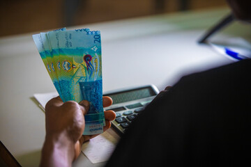 Dark skinned African person in black top working on desk holding Kuwaiti Dinar notes in hand and pressing punching calculator with receipt on table. accounting concept