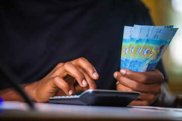 Dark skinned African person in black top working on desk holding Kuwaiti Dinar notes in hand and pressing punching calculator with receipt on table.  accounting concept