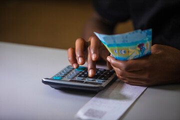 Dark skinned African person in black top working on desk holding Kuwaiti Dinar notes in hand and pressing punching calculator with receipt on table.  accounting concept