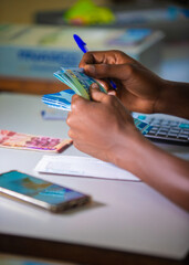 vertical shot of Dark skinned African person working on desk holding counting Ghanaian cedi notes money with some notes on table. Top view accounting concept