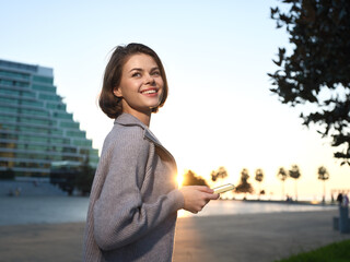 A cheerful woman stands outdoors during sunset, holding a smartphone. She smiles warmly, wearing a light coat, projecting confidence and optimism in a modern urban setting.